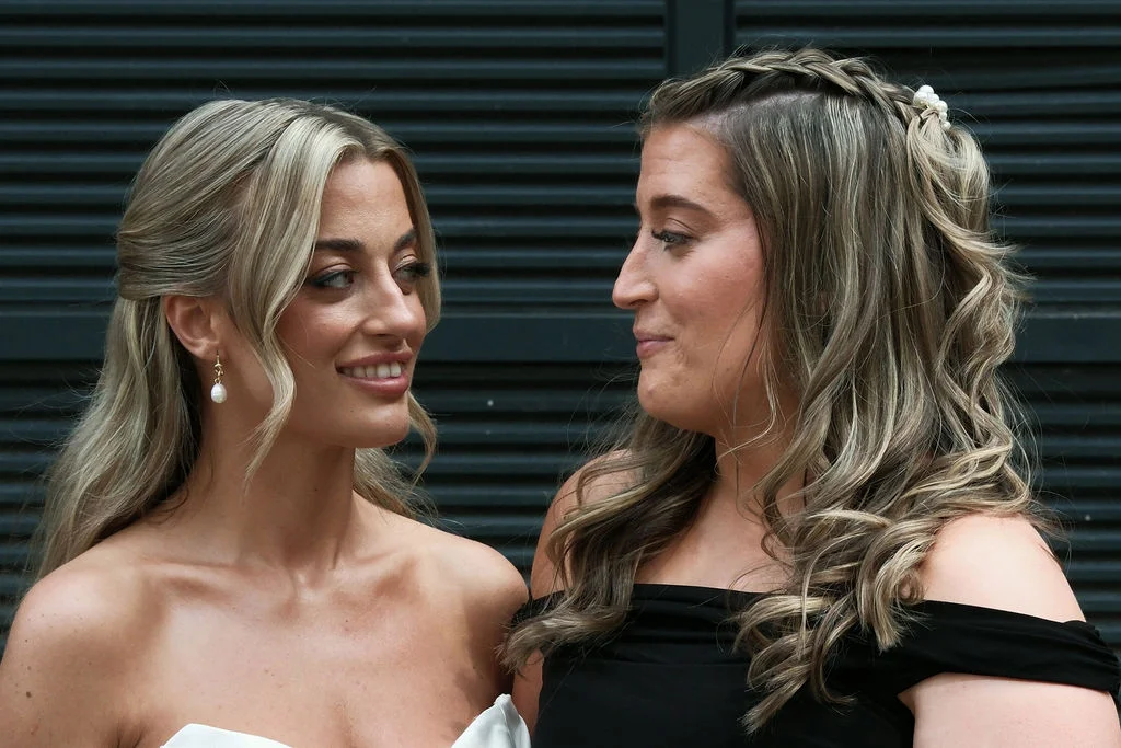 Two smiling brides share a tender glance outdoors against a dark slatted background, showcasing elegant half-up hairstyles and statement pearl earrings.