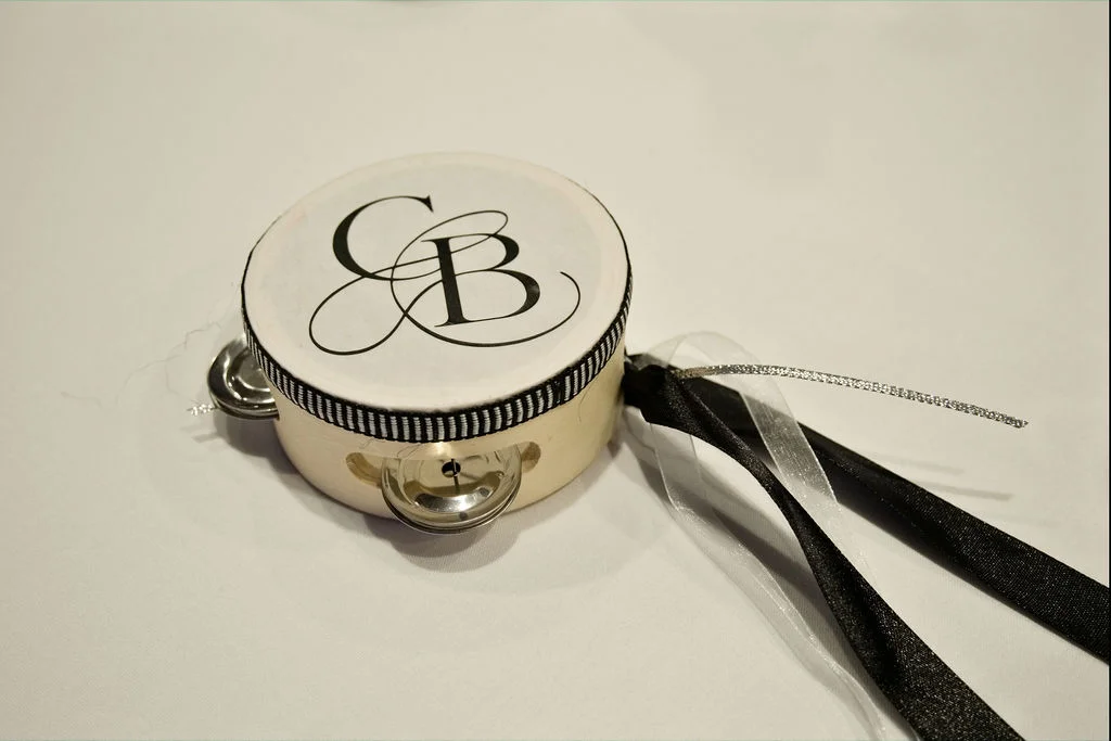 A personalised white and black tambourine wedding favour rests on a plain surface, decorated with a monogram and black and silver ribbons.