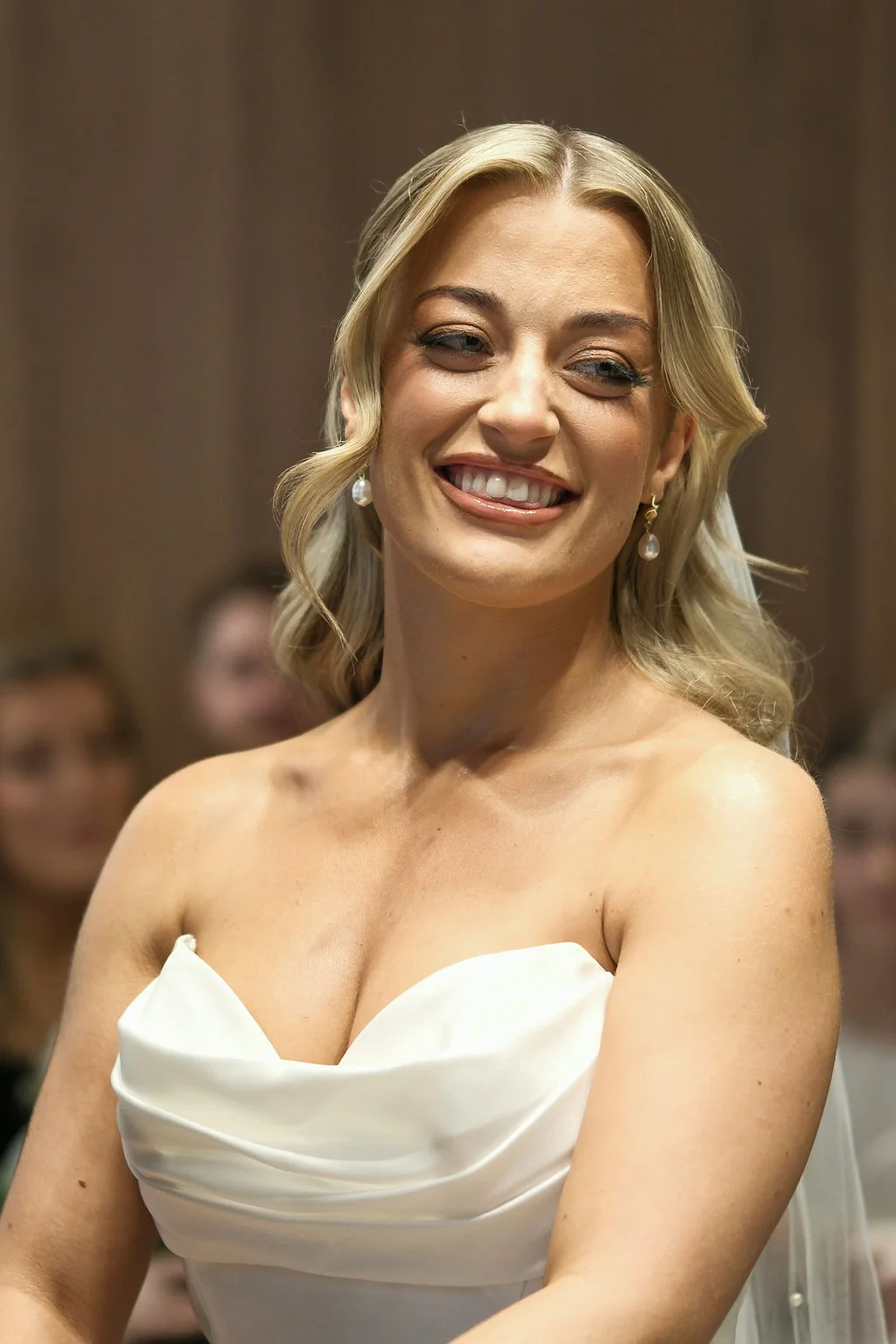 A stunning bride with blonde hair styled half up and down smiles radiantly in a strapless satin wedding dress featuring a draped sweetheart neckline.
