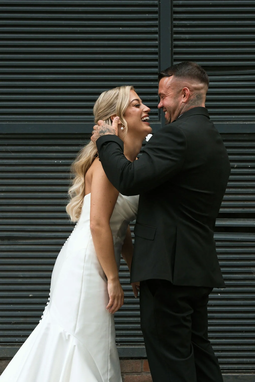 A newlywed couple shares a joyful laughing moment against a dark slatted background, with the bride wearing a sleek satin gown and the groom in a classic black suit.