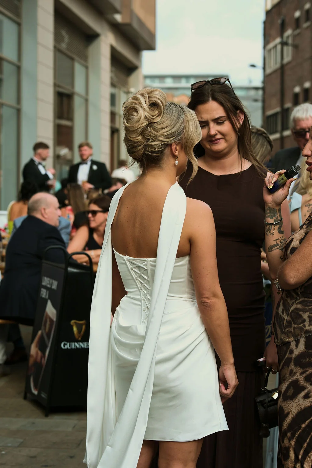 A bride with an elaborate updo and a short satin dress with a lace-up back stands outside a city venue chatting with guests.
