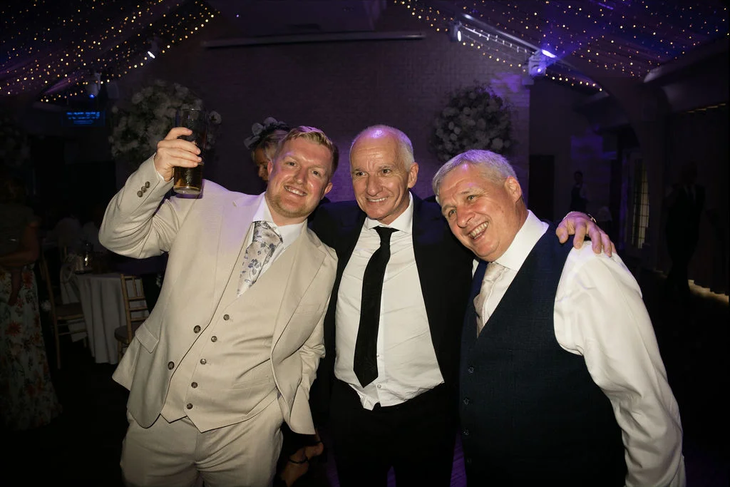 Three smiling male wedding guests pose together during the evening reception beneath a canopy of glowing fairy lights.