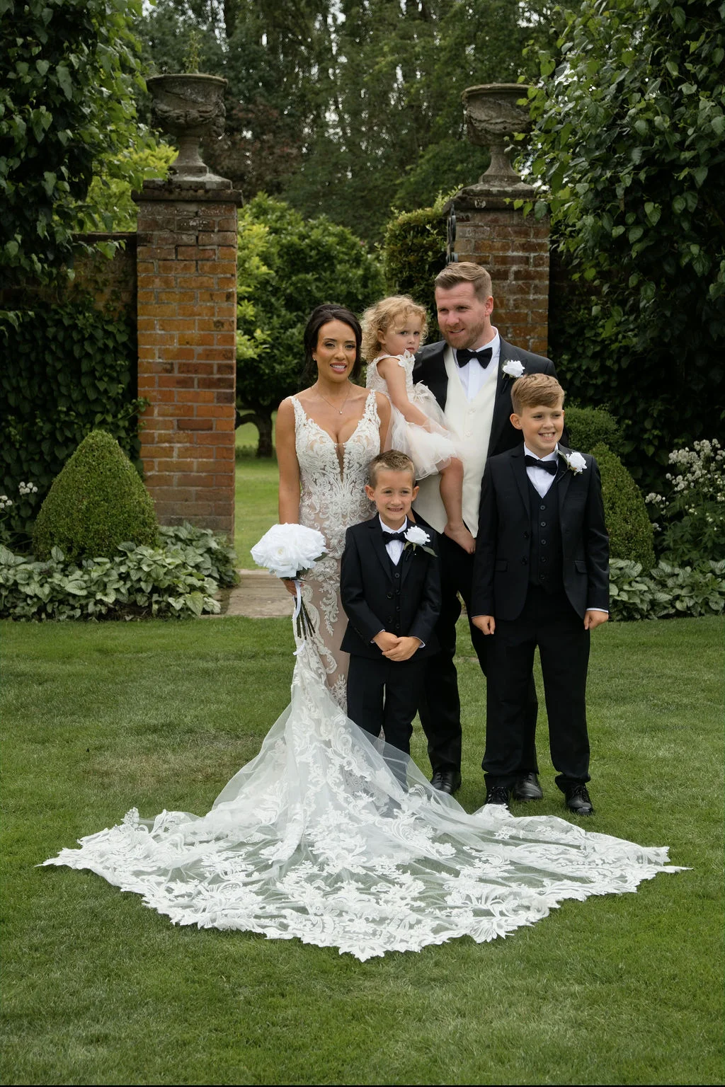 A newlywed couple poses in a formal group portrait on a manicured lawn with their young children outside a historic brick walled garden.