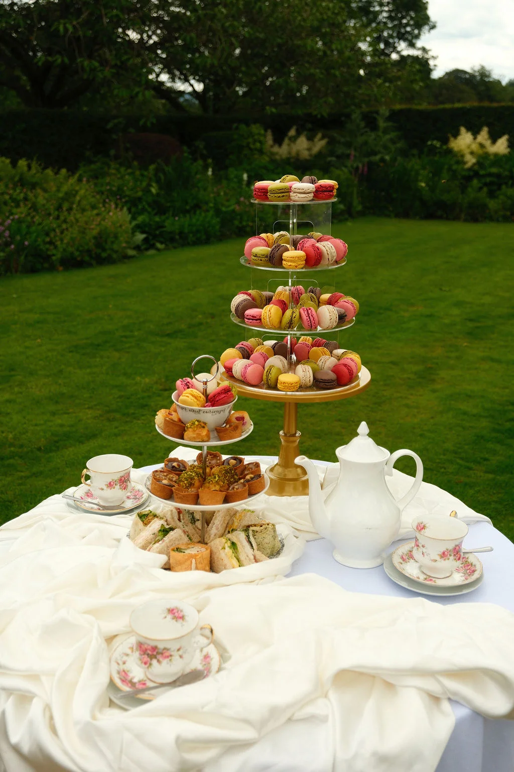 An outdoor afternoon tea setup on a lush green lawn features a tiered macaron tower, dainty sandwiches, and vintage floral teacups on a white linen tablecloth.