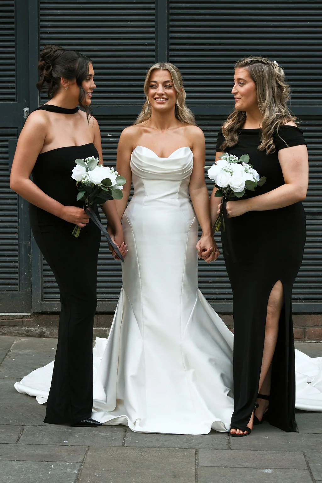 A smiling bride in a strapless white satin gown holds hands with two bridesmaids wearing matching black dresses and holding white rose bouquets outside a dark slatted venue.