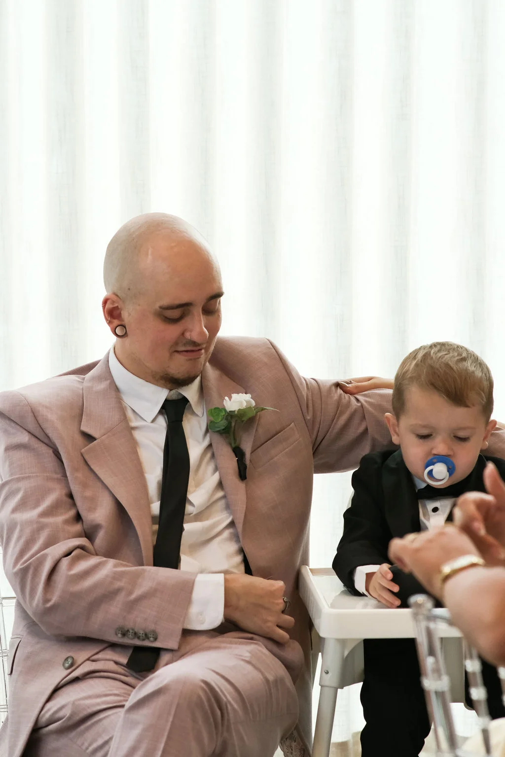 A groom wearing a pink suit with a black tie sits next to a young child in a high chair wearing a miniature tuxedo.