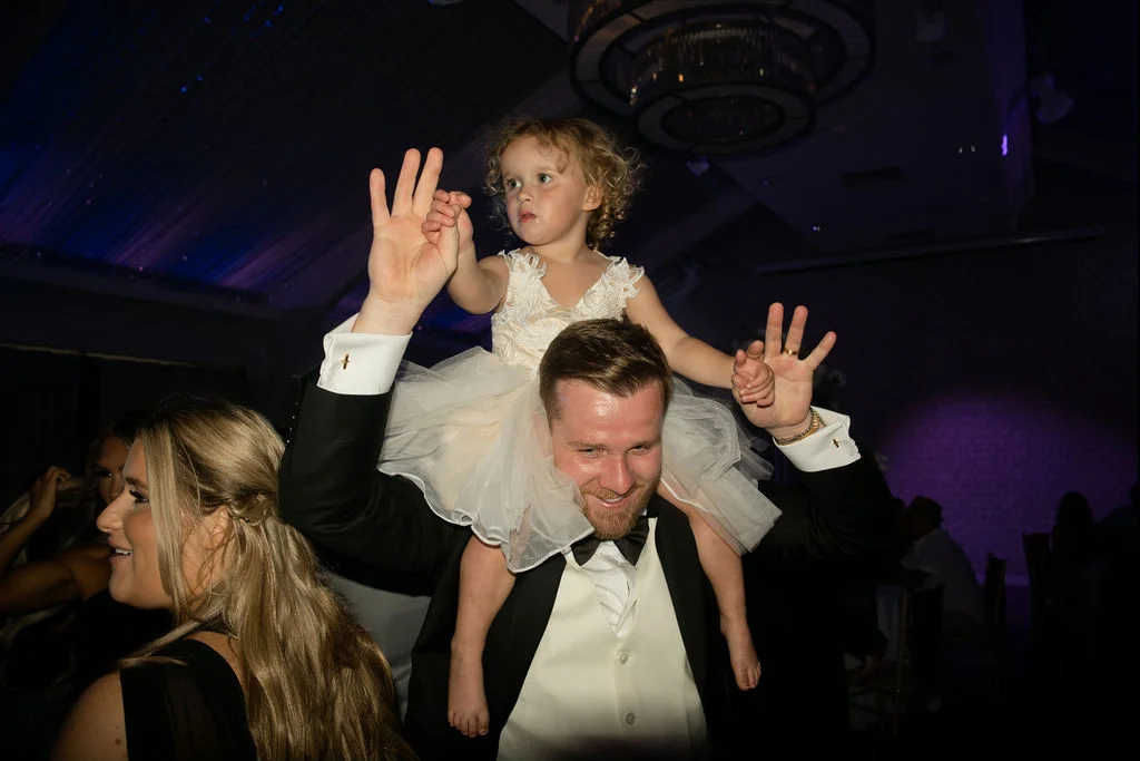 A young flower girl in a white tulle dress rides happily on the shoulders of a man wearing a black tuxedo and white waistcoat on a lively wedding dance floor.