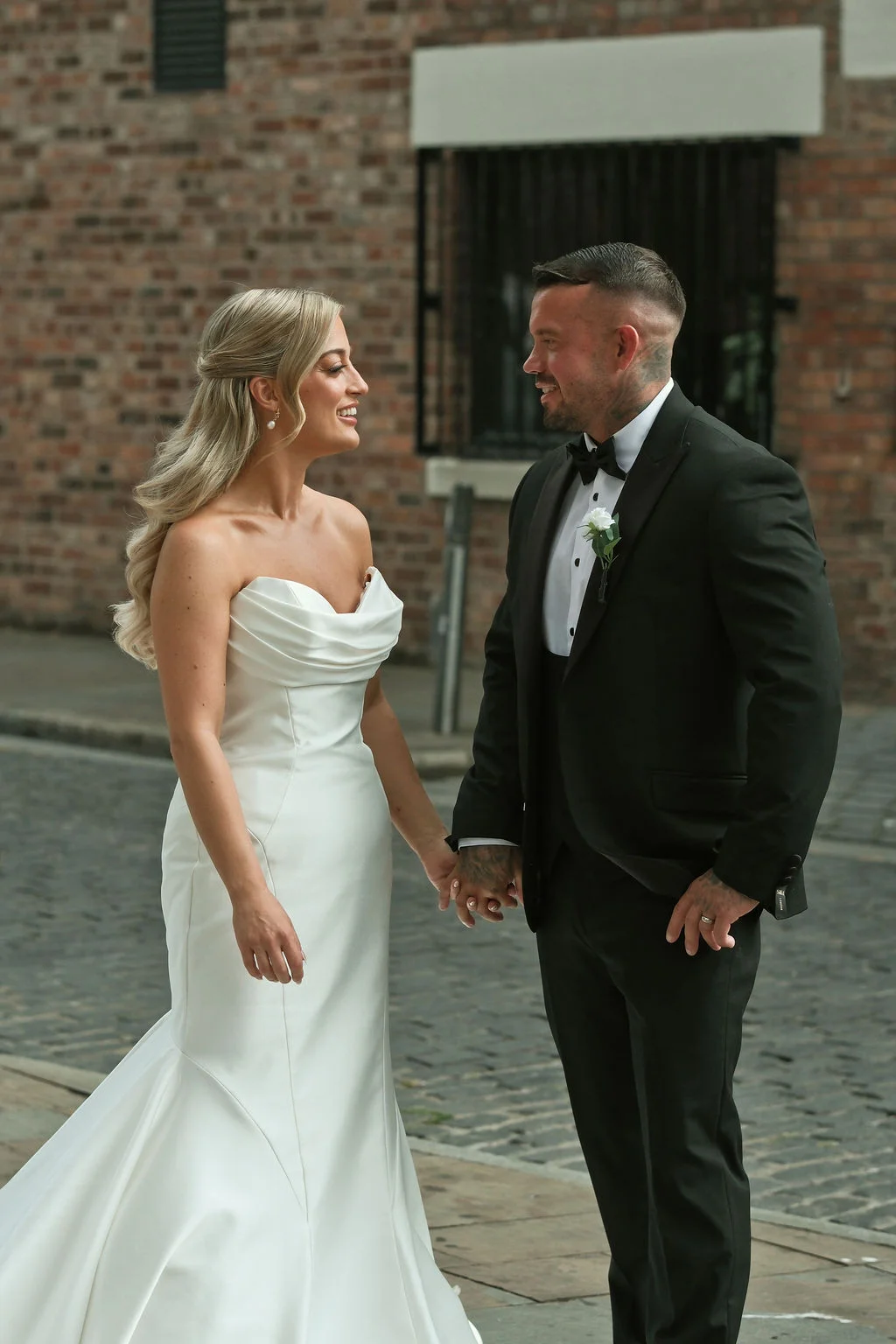 A newlywed couple holds hands and smiles at each other on a cobbled city street outside a brick building.