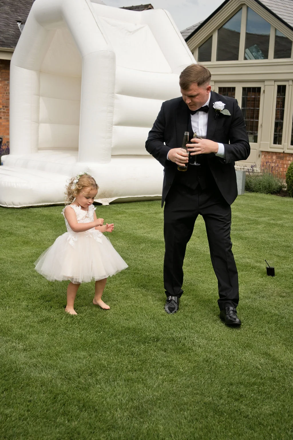 A flower girl in a white dress dances barefoot on the lawn next to a groom holding drinks in front of a white bouncy castle.