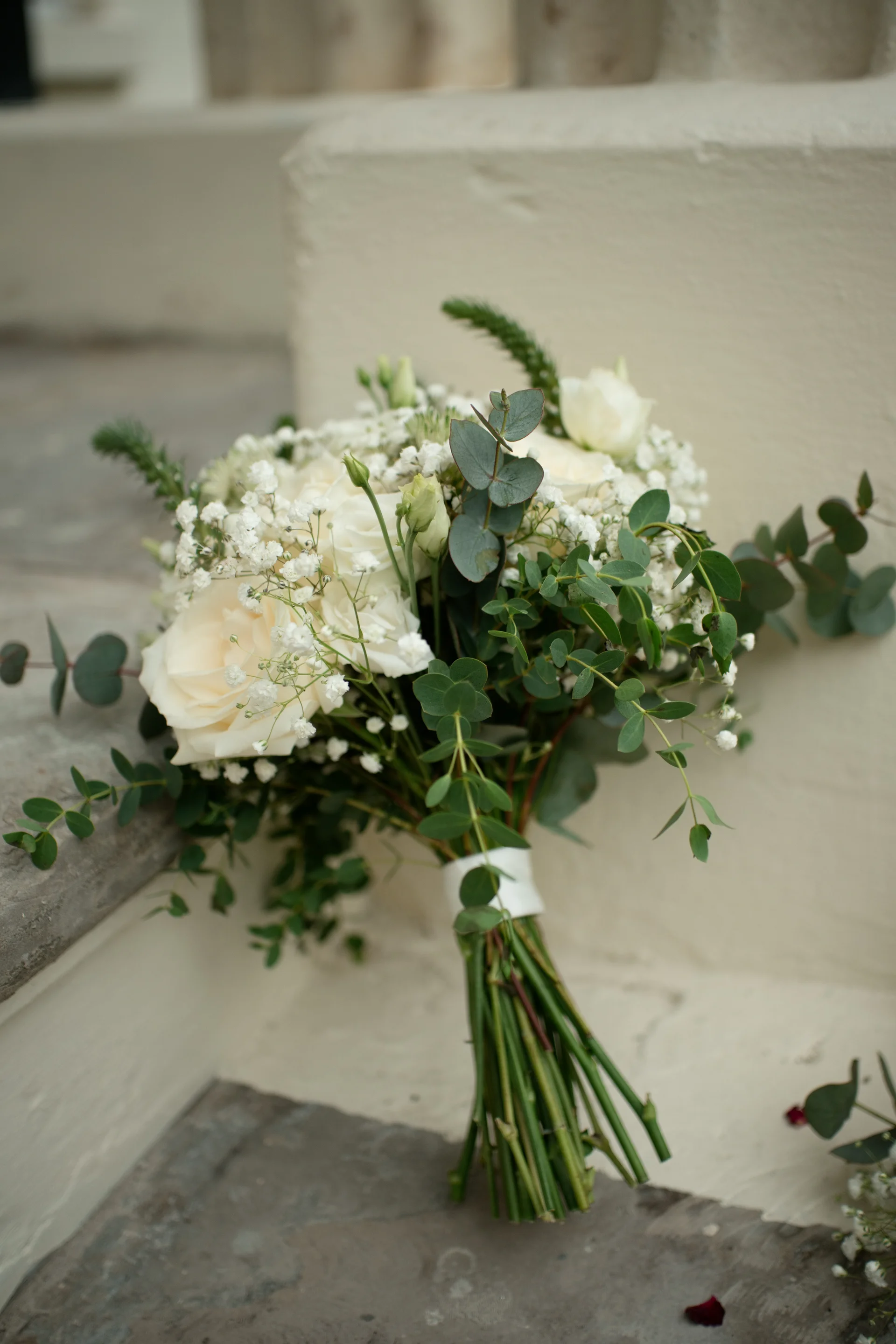 An elegant hand-tied bridal bouquet featuring white roses, delicate baby's breath, and trailing eucalyptus rests on a stone step.