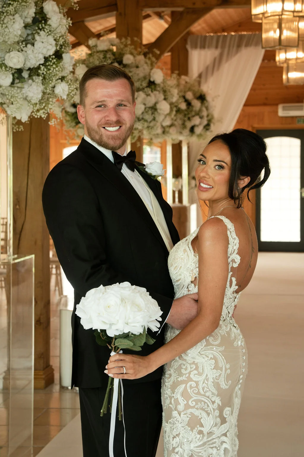 A smiling bride and groom pose inside a rustic oak-framed barn decorated with white floral installations, with the bride holding a white rose bouquet and wearing a lace gown.