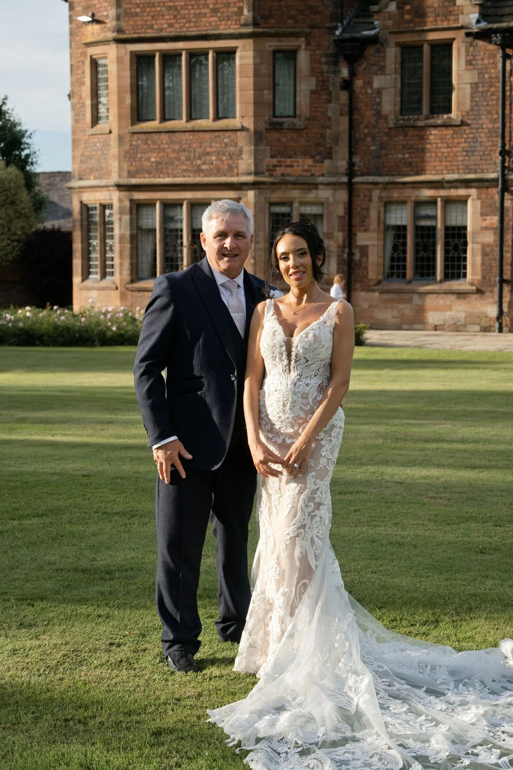 A smiling bride in an intricate lace gown stands proudly with her father on the manicured lawn of a grand historic manor house.
