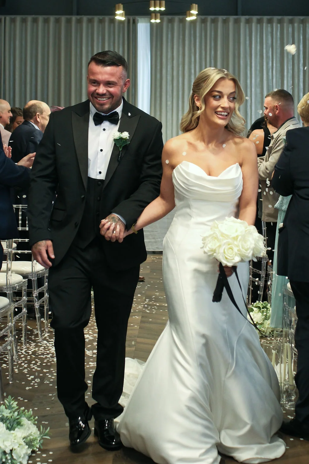 A glowing newly married couple walk hand-in-hand down the aisle amidst a shower of white confetti, smiling brightly as guests cheer them on in a contemporary venue.