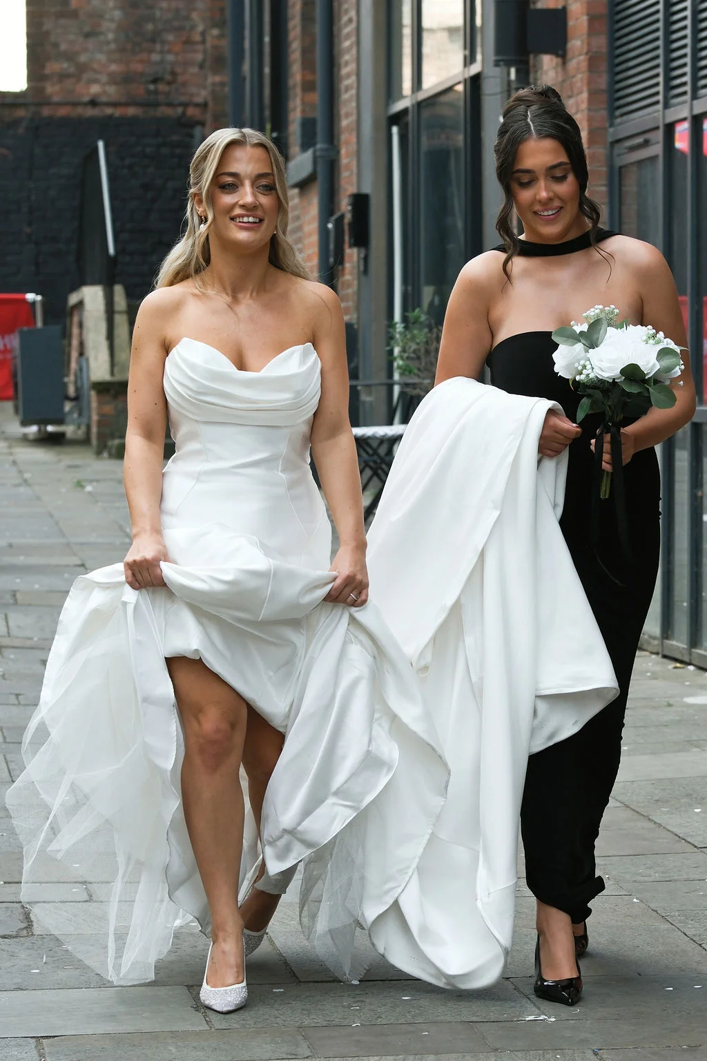 A smiling bride wearing a strapless satin gown with a thigh split walks down a paved urban street alongside a bridesmaid in a black dress carrying a bouquet.