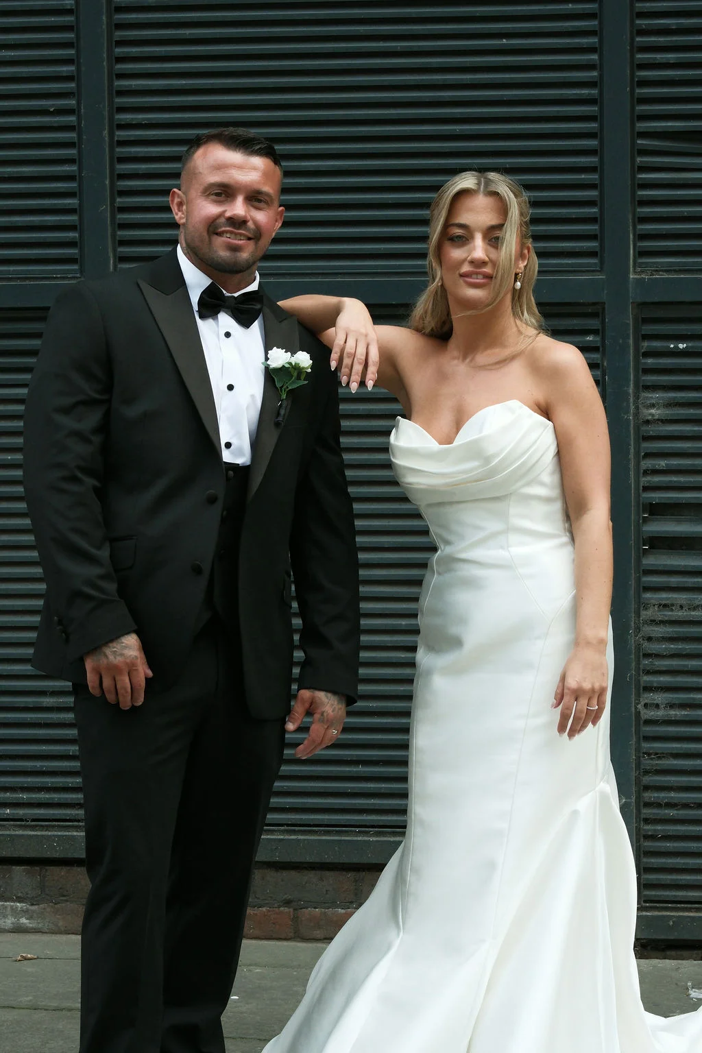 A newlywed couple poses together against a dark slatted backdrop, with the groom in a classic black tuxedo and the bride in an elegant strapless satin gown.