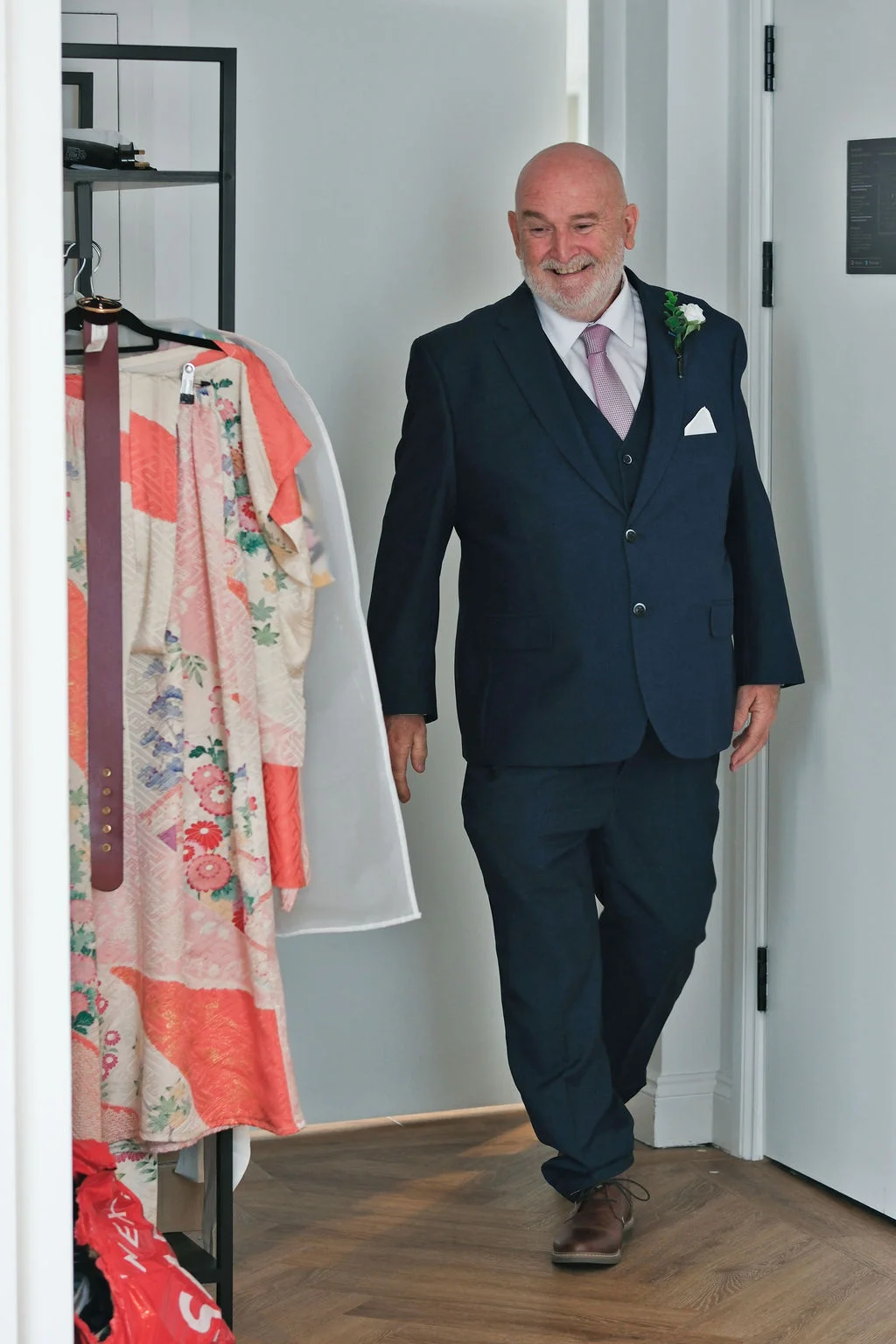A smiling, mature father of the bride wearing a navy three-piece suit and a buttonhole walks through a doorway during wedding morning preparations.