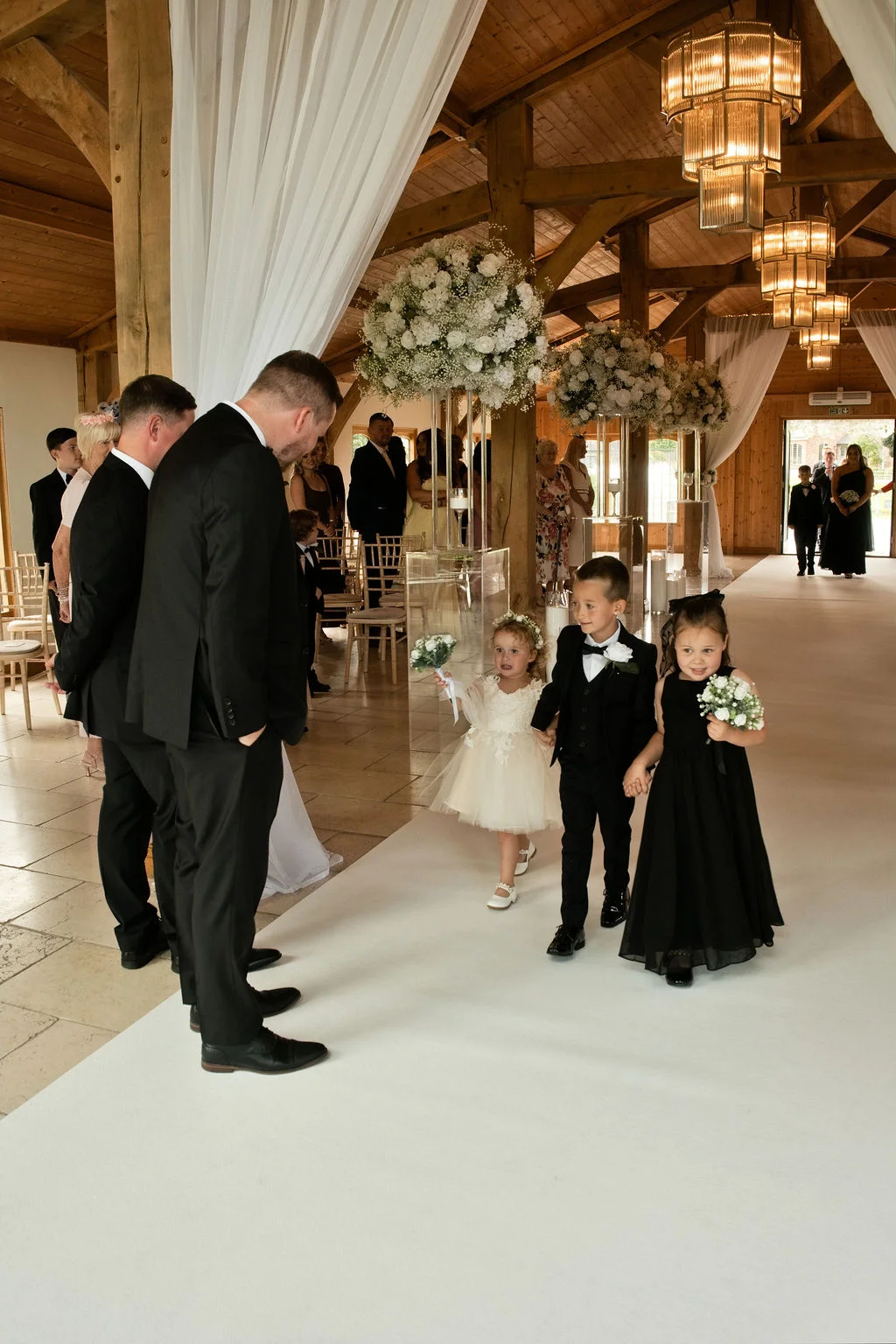 Young flower girls and a page boy walk down the aisle in a rustic oak-framed barn decorated with white floral pedestals and elegant chandeliers.