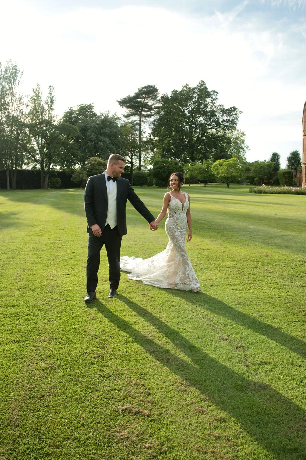 A newlywed couple walks hand in hand across a manicured lawn during golden hour, casting long shadows behind them.