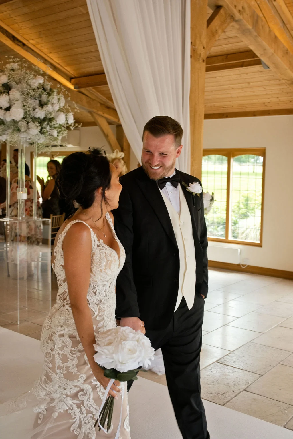A smiling bride and groom walk hand in hand down the aisle inside an oak framed barn venue, with the bride holding a white rose bouquet.