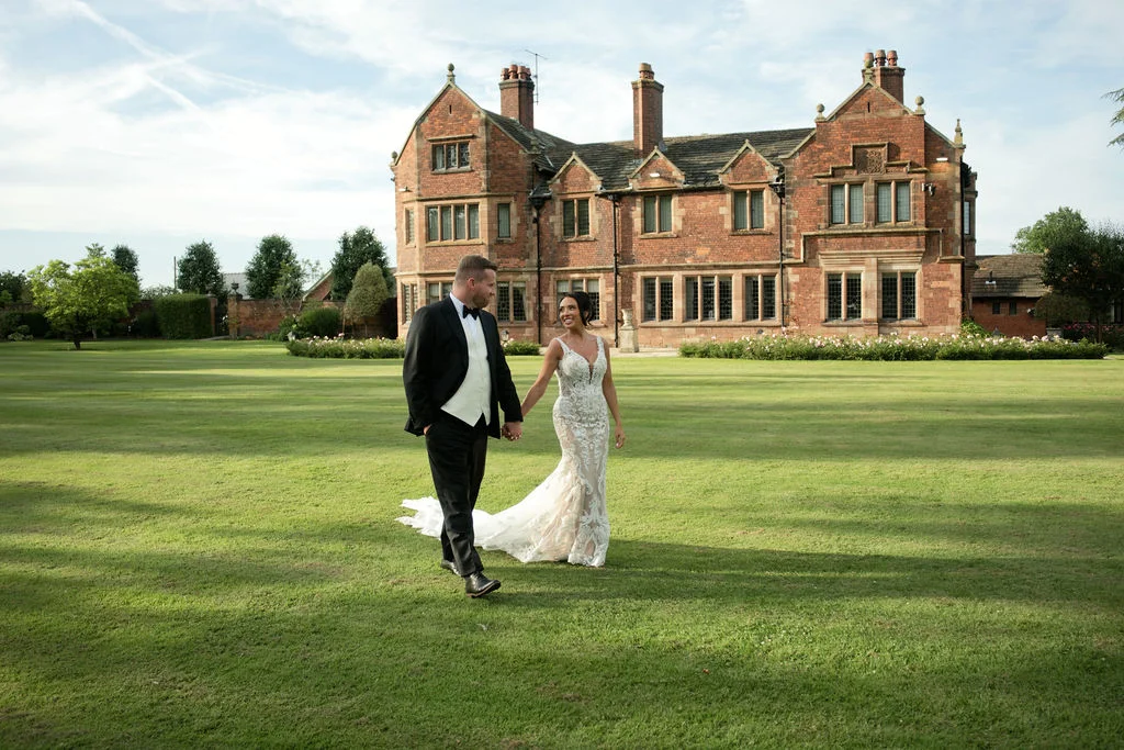 A newlywed couple walk hand in hand across a vast green lawn in front of a grand red brick manor house.