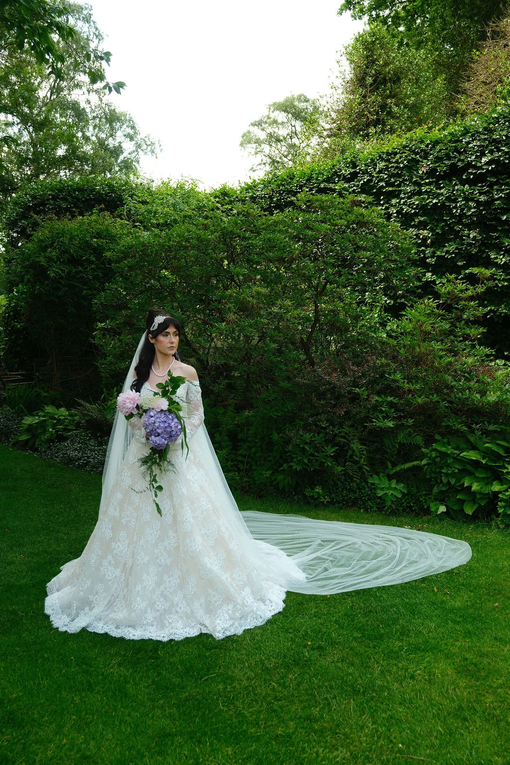 A stunning bride poses in a lush green garden wearing an off-the-shoulder lace a-line wedding dress, holding a cascading bouquet of pastel hydrangeas with a long veil pooling on the lawn.