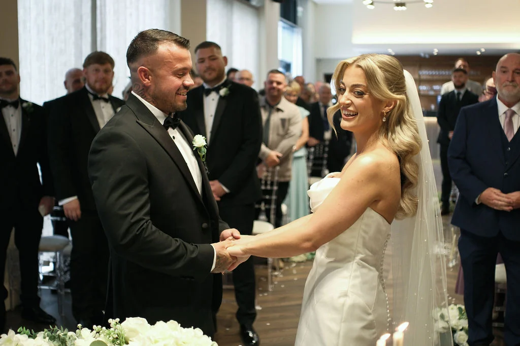 A joyful bride and groom hold hands and smile at each other during their indoor wedding ceremony, surrounded by family and friends.