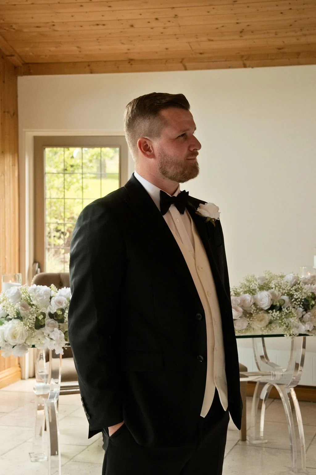 A groom stands proudly in a black tuxedo with a cream waistcoat and a buttonhole flower, captured in a bright indoor setting.