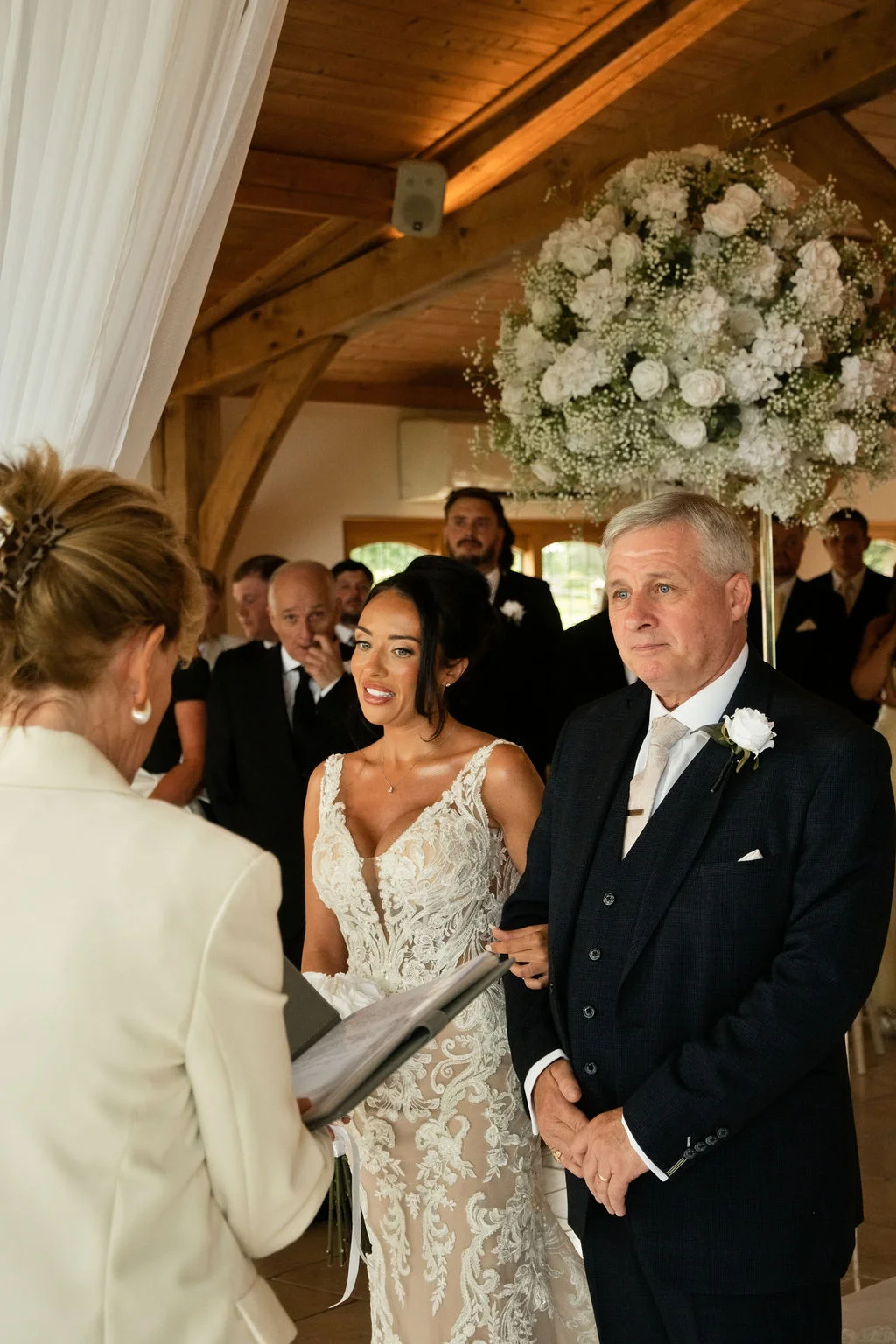 A bride walking down the aisle with her father during an indoor wedding ceremony in a rustic barn venue decorated with white floral arrangements.