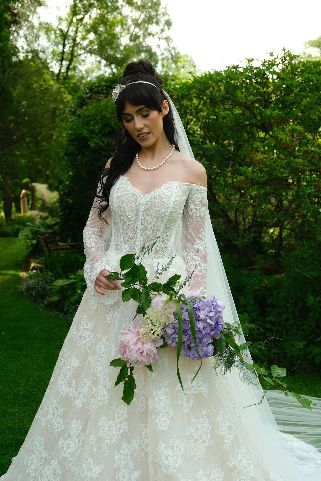 A dark-haired bride in an off-the-shoulder lace gown with long sleeves and a corset bodice stands outdoors holding a cascading bouquet of pink peonies and purple hydrangeas.