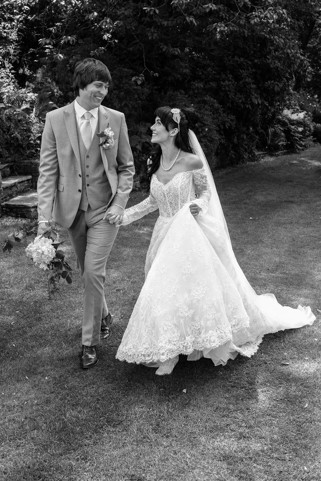 A bride and groom smile at each other while walking hand in hand through a lush garden on their wedding day.