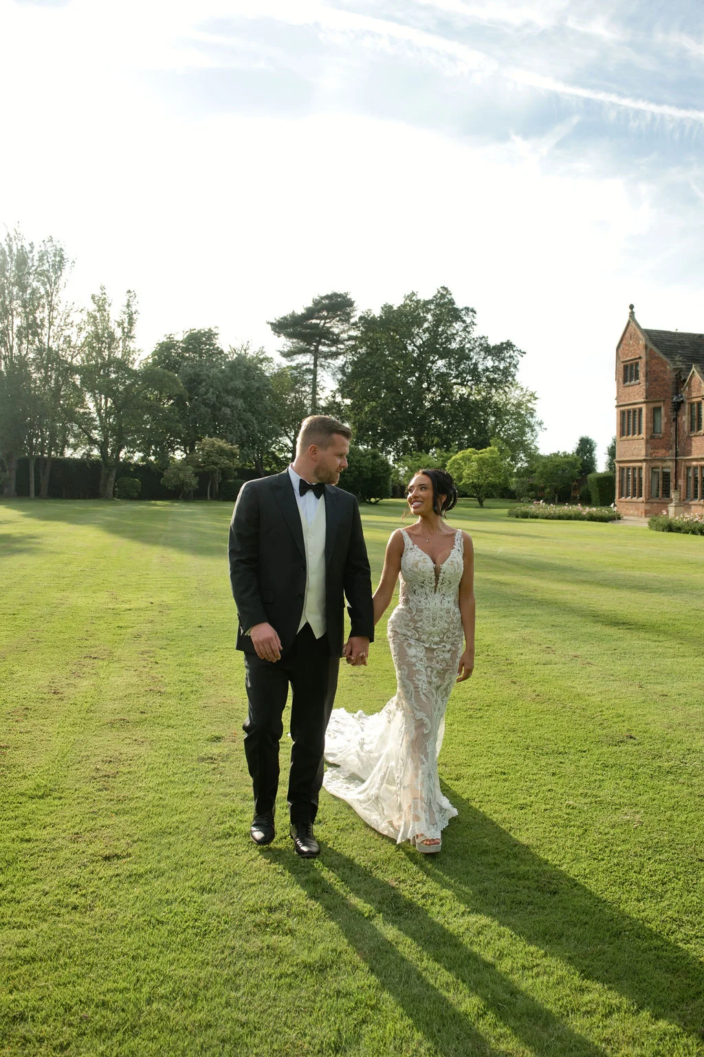 Newlyweds walk hand in hand across the manicured lawn of a stately manor house in the golden afternoon sunshine.