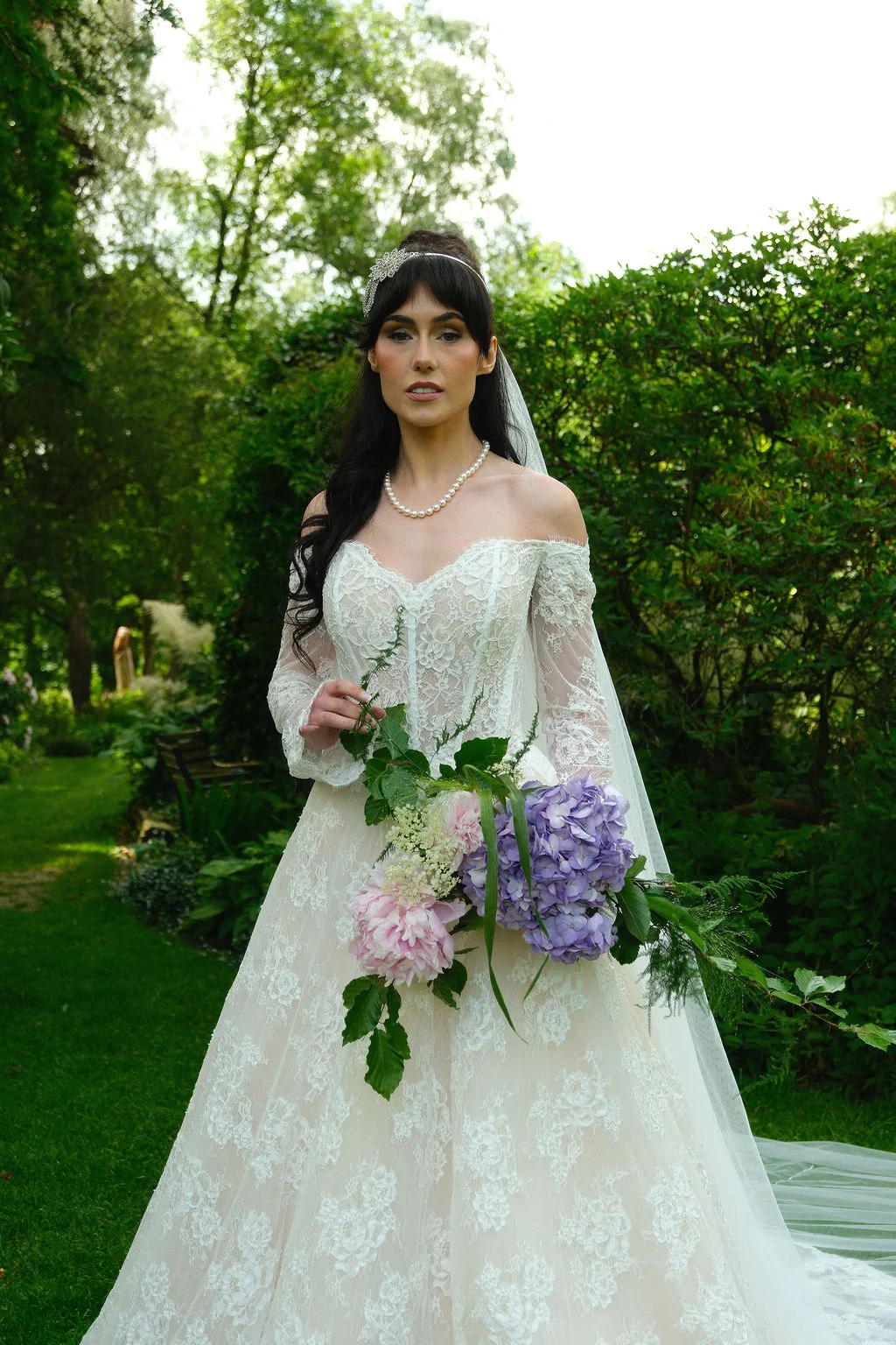 A stunning bride stands in a lush garden setting wearing an off-the-shoulder lace wedding dress and holding a cascading bouquet of purple hydrangeas and pink peonies.