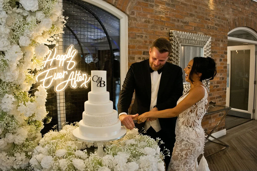 A joyful bride and groom laugh together while cutting a multi-tiered white wedding cake surrounded by lush floral arrangements and a glowing neon sign against an exposed brick wall.