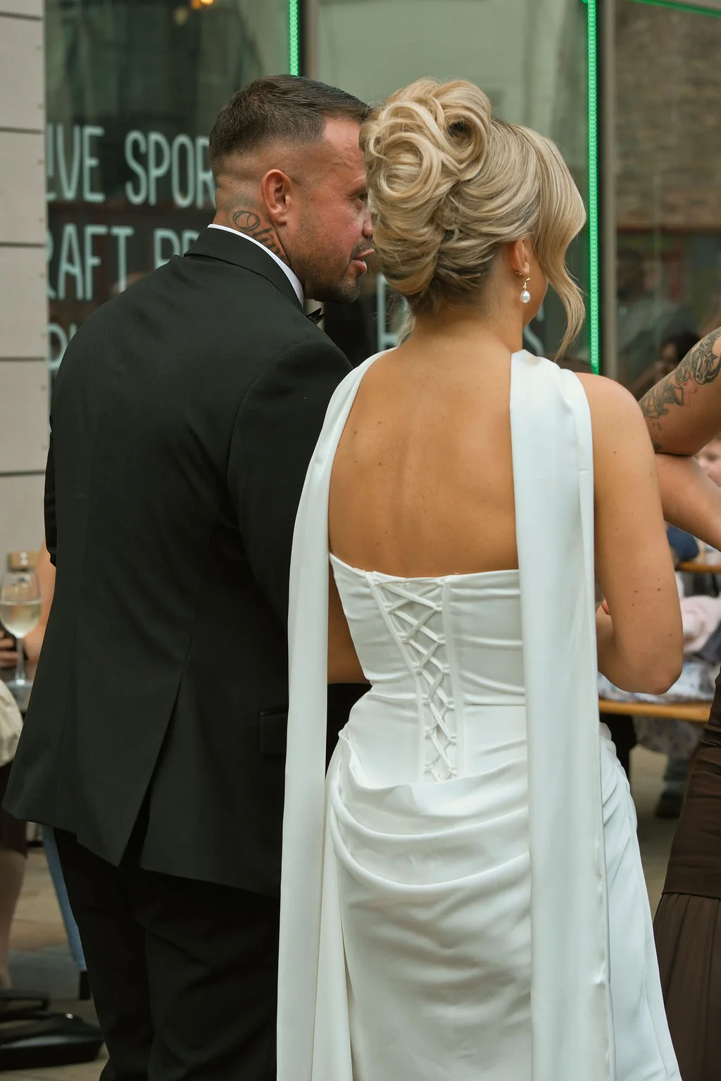 A newlywed couple is viewed from behind as they walk together outdoors, featuring the bride in a satin gown with a lace-up back and cape, alongside the groom in a classic black suit.