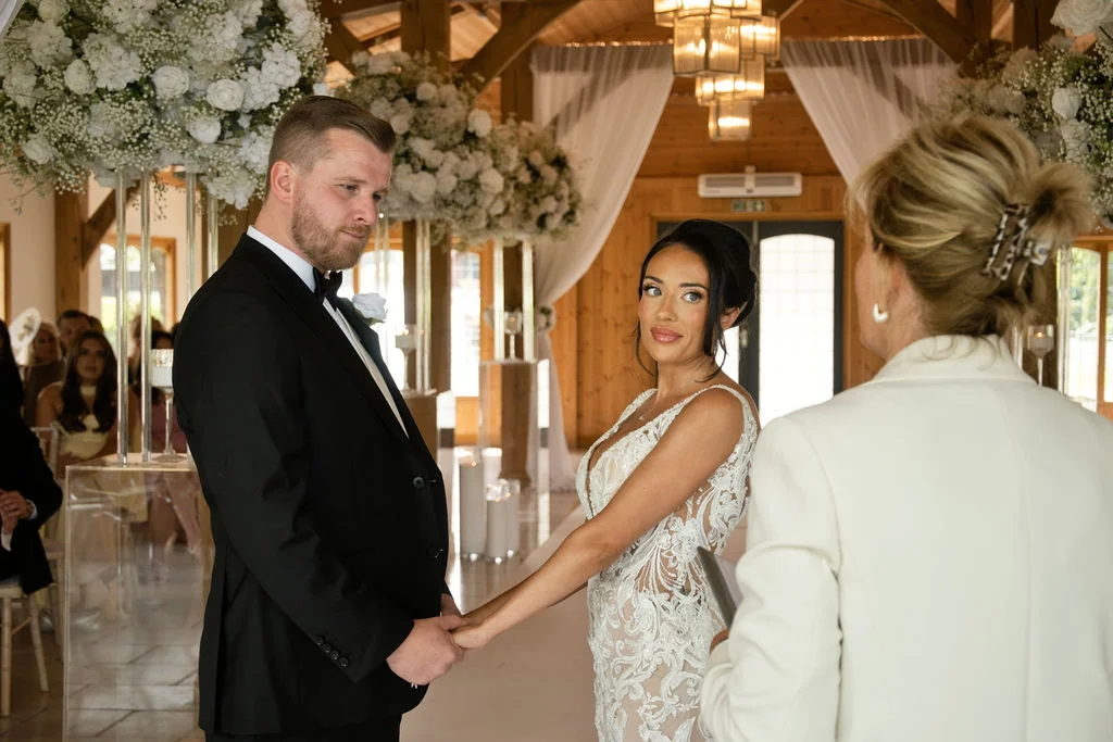 The bride and groom hold hands and exchange glances during their civil ceremony inside an oak-framed barn decorated with lavish white floral arrangements.
