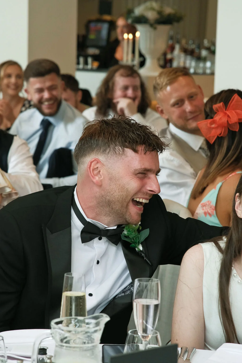 A guest in a black tuxedo and bow tie shares a hearty laugh during the wedding reception speeches.