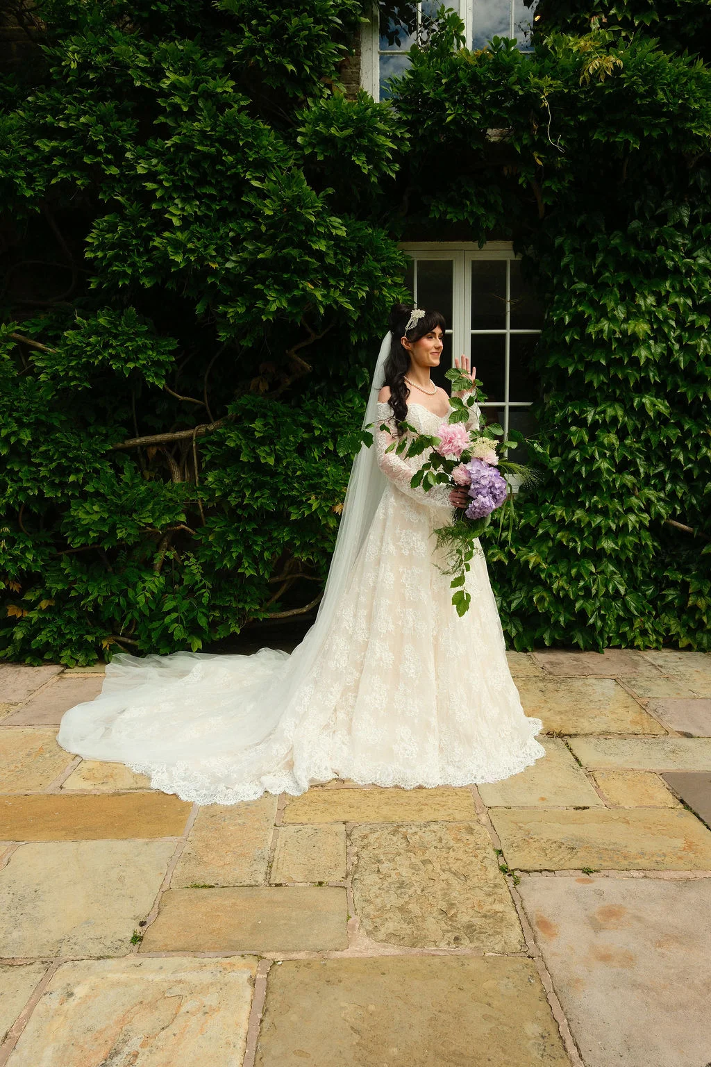 A stunning bride poses in a lace off-the-shoulder gown with a long train and veil, holding a cascading bouquet of pink and purple blooms against an ivy-clad wall.