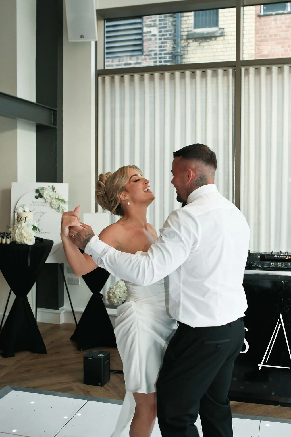 A newlywed couple share a joyful first dance together on an illuminated dance floor inside a modern industrial wedding venue.