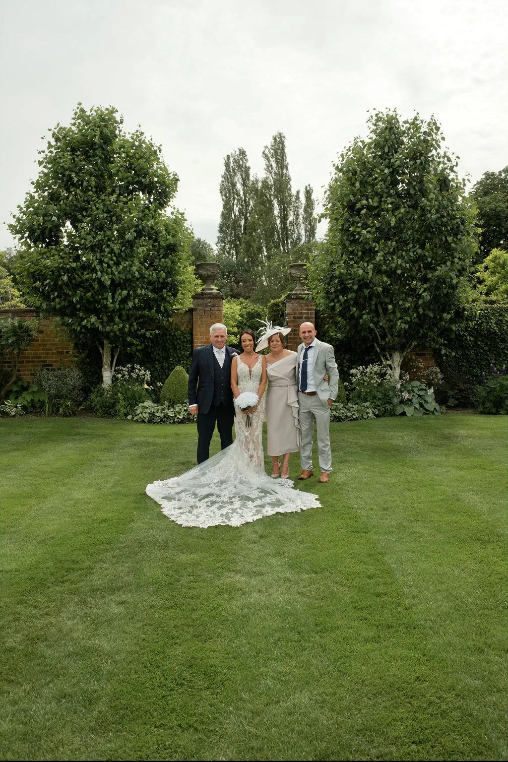 A bridal party comprising a bride, groom, and parents poses on a manicured lawn in front of tall trees and a walled garden backdrop.