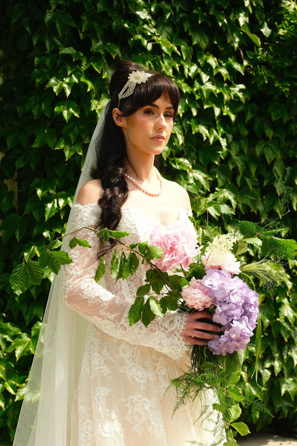 A dark-haired bride wearing an off-the-shoulder lace wedding dress and a pearl necklace poses outdoors holding a cascading bouquet of pink peonies and purple hydrangeas in front of a lush ivy wall.