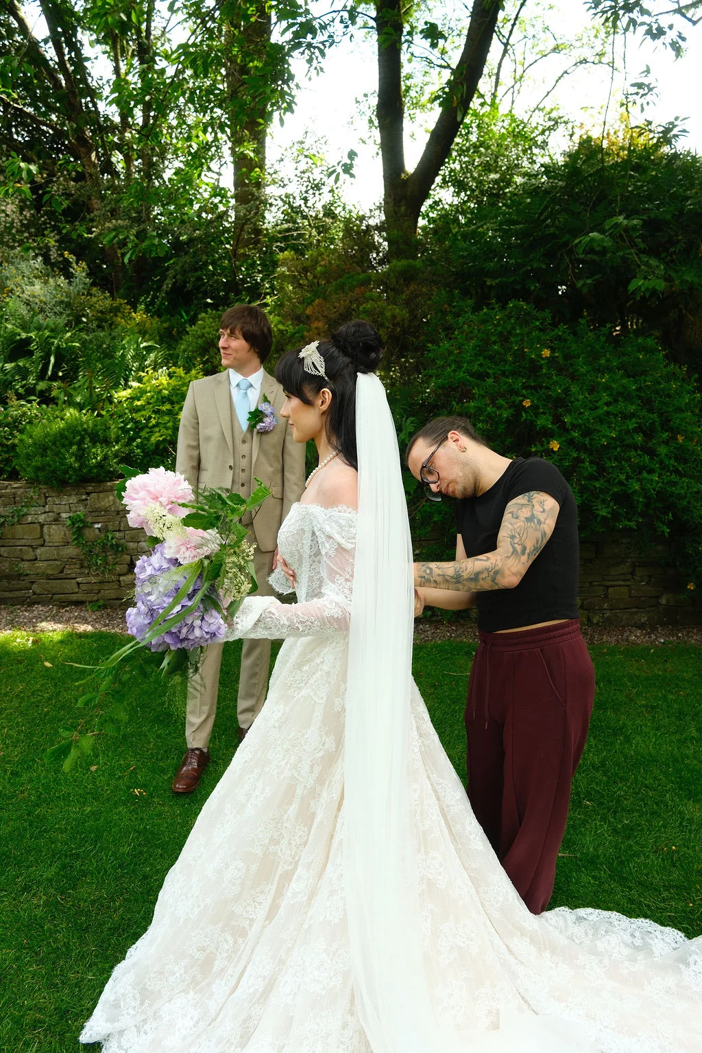 A bride in an off-the-shoulder lace gown with a long veil receives final adjustments in a lush garden setting while the groom looks on.