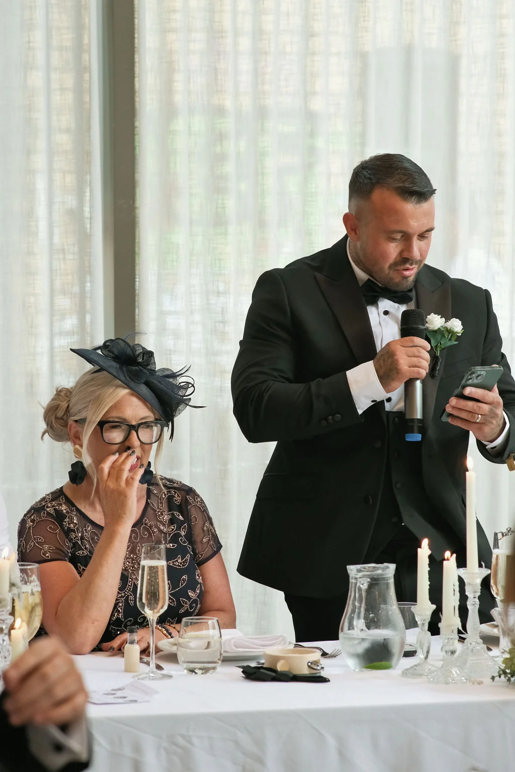 A groom in a black tuxedo delivers a speech holding a microphone and mobile phone, while a seated woman in a dark patterned dress and fascinator reacts emotionally.
