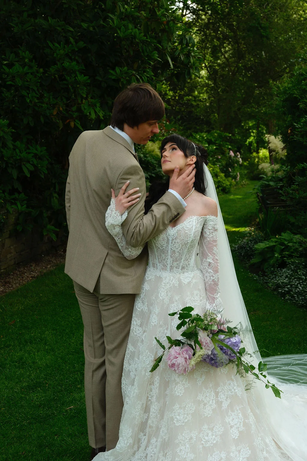 A newlywed couple embraces tenderly in a lush green garden, with the bride looking up at the groom while holding a soft floral bouquet.