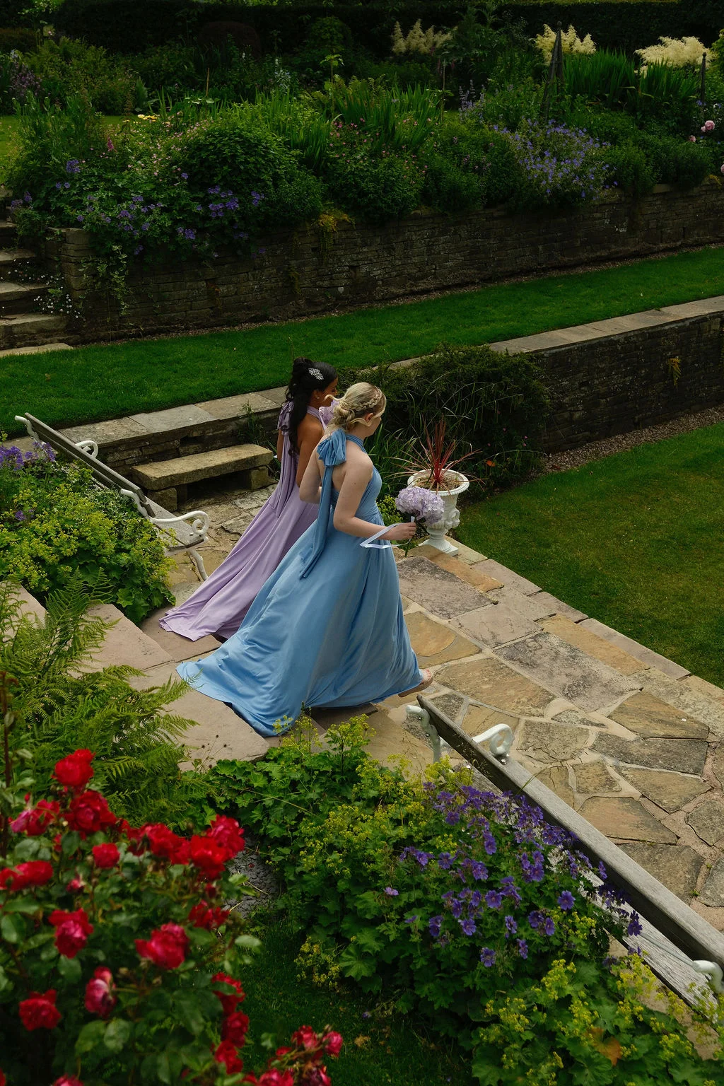 Two bridesmaids walk down stone garden steps carrying blue and purple bouquets in a manicured English country garden.