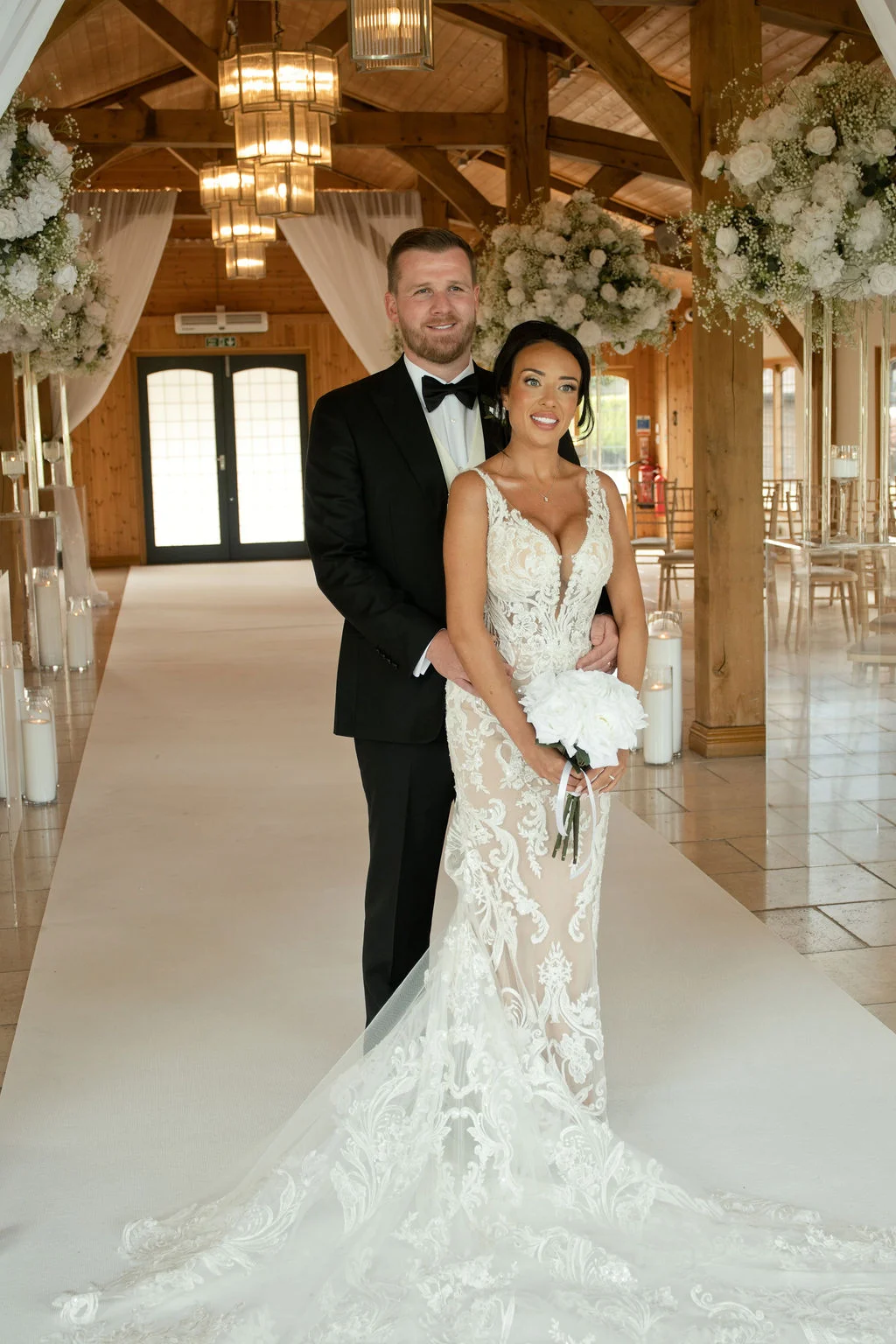 A newlywed couple poses inside an oak framed barn featuring exposed rafters and a vaulted ceiling, with the bride holding a white bouquet while wearing a fitted lace wedding dress with a long train, and the groom wearing a classic black tuxedo.