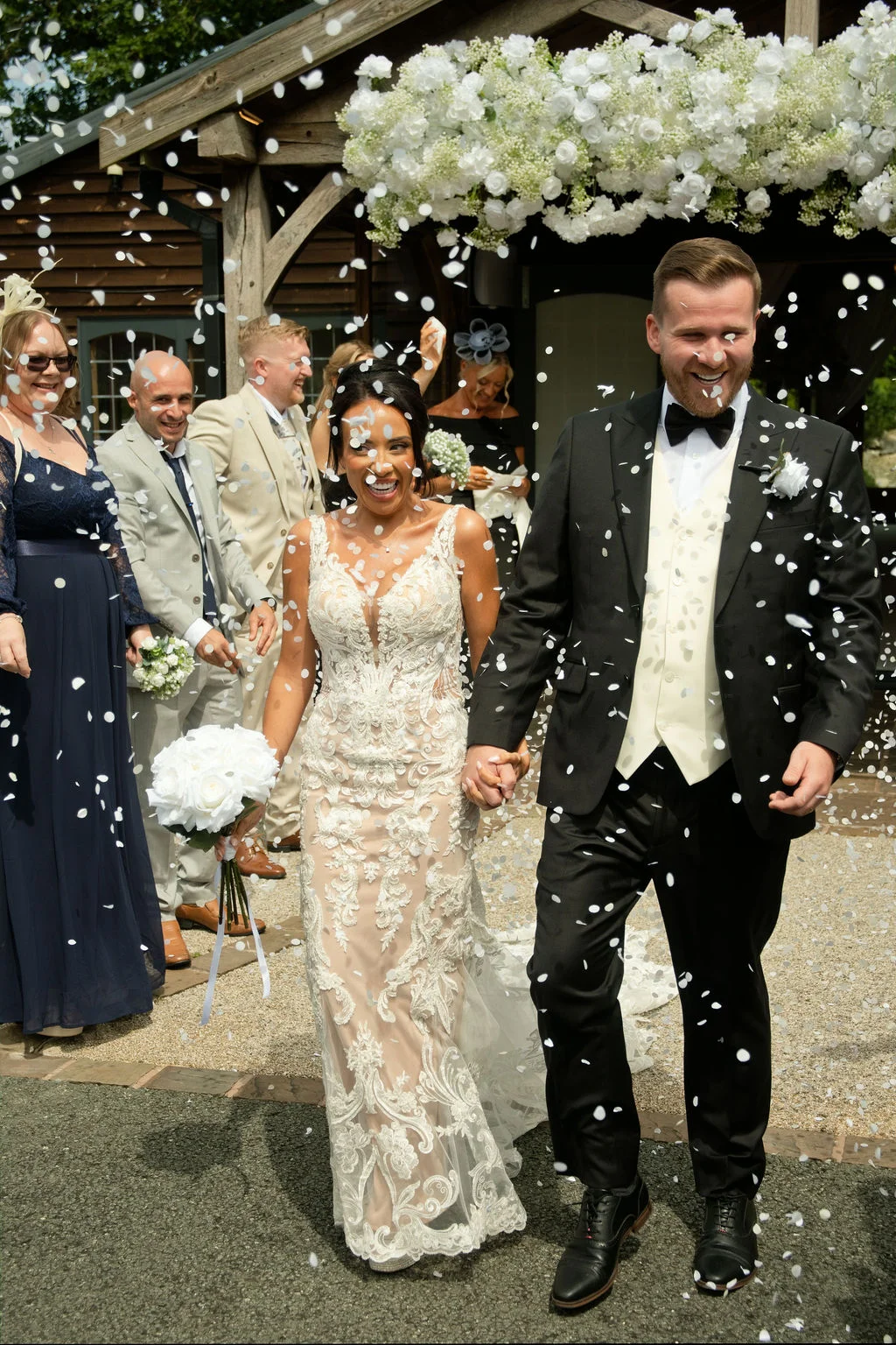 A joyful newlywed couple walks hand in hand through a shower of white confetti outside an oak-framed barn decorated with a lush floral arch.