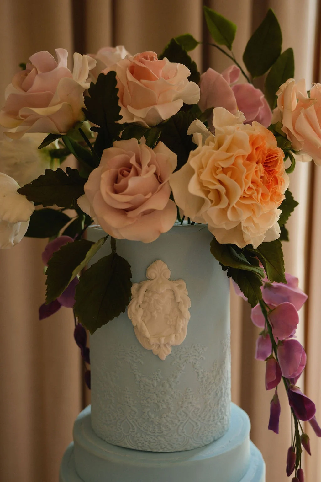 A close-up of a tiered blue wedding cake adorned with delicate white lace detailing, an intricate white cameo emblem, and an elaborate arrangement of sugar roses and pastel flowers.