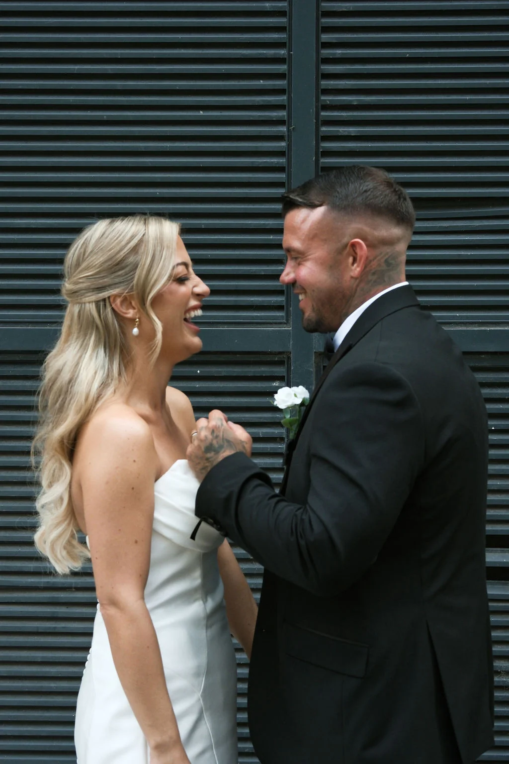 A newlywed couple shares a joyful laughing moment together outdoors against a dark slatted background, with the bride wearing a strapless white gown and the groom in a classic black suit.
