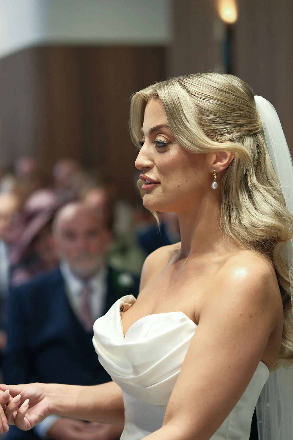 A profile portrait of a blonde bride with half-up wavy hair wearing a strapless satin gown and pearl earrings during a wedding ceremony.