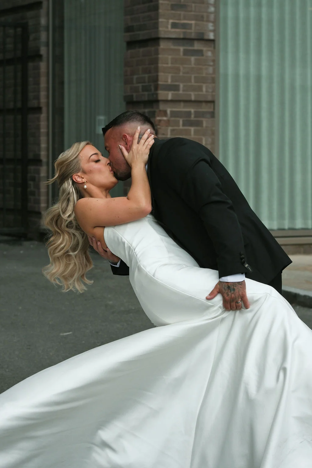 A newlywed couple shares a dramatic dip kiss in front of a modern urban building with brick detailing.